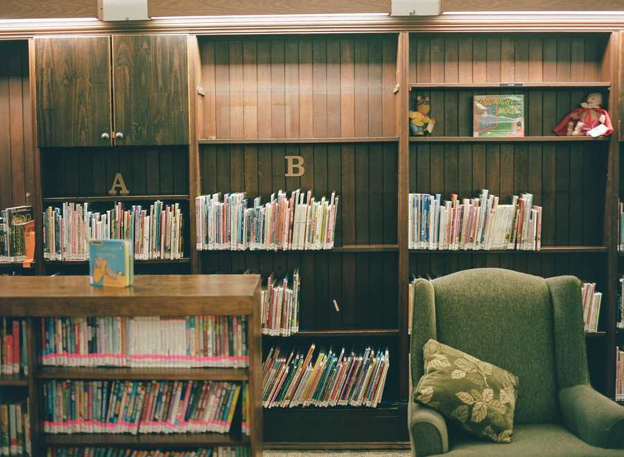 This is the top left portion of a composite of four images that make up one image of a green chair in a library. This section contains the upper left portion of the chair, which sits next to a bookshelf filled with books. Behind the chair is a wall of bookshelves filled with colorful children’s books, labeled A and B in this portion of the image.
