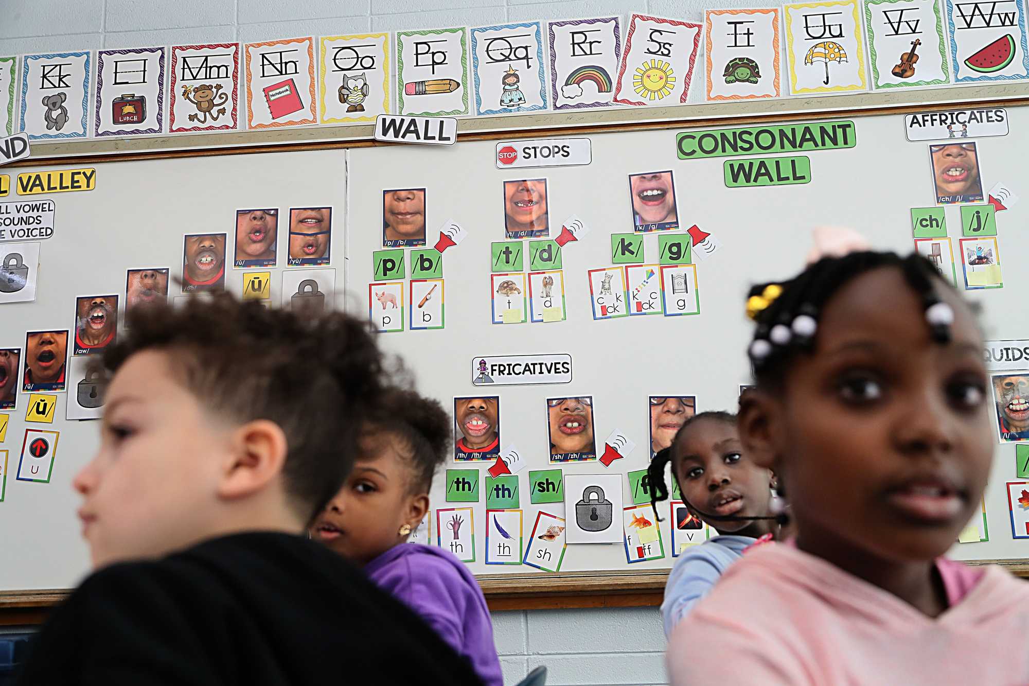 First grade students at Margaret L. Donovan Elementary School in Randolph listened to their teacher during a reading lesson. A “sound wall” on the white board behind them shows students how to form different sounds with their mouths. Randolph school leaders say a new structured literacy curriculum has helped improve students' reading skills.