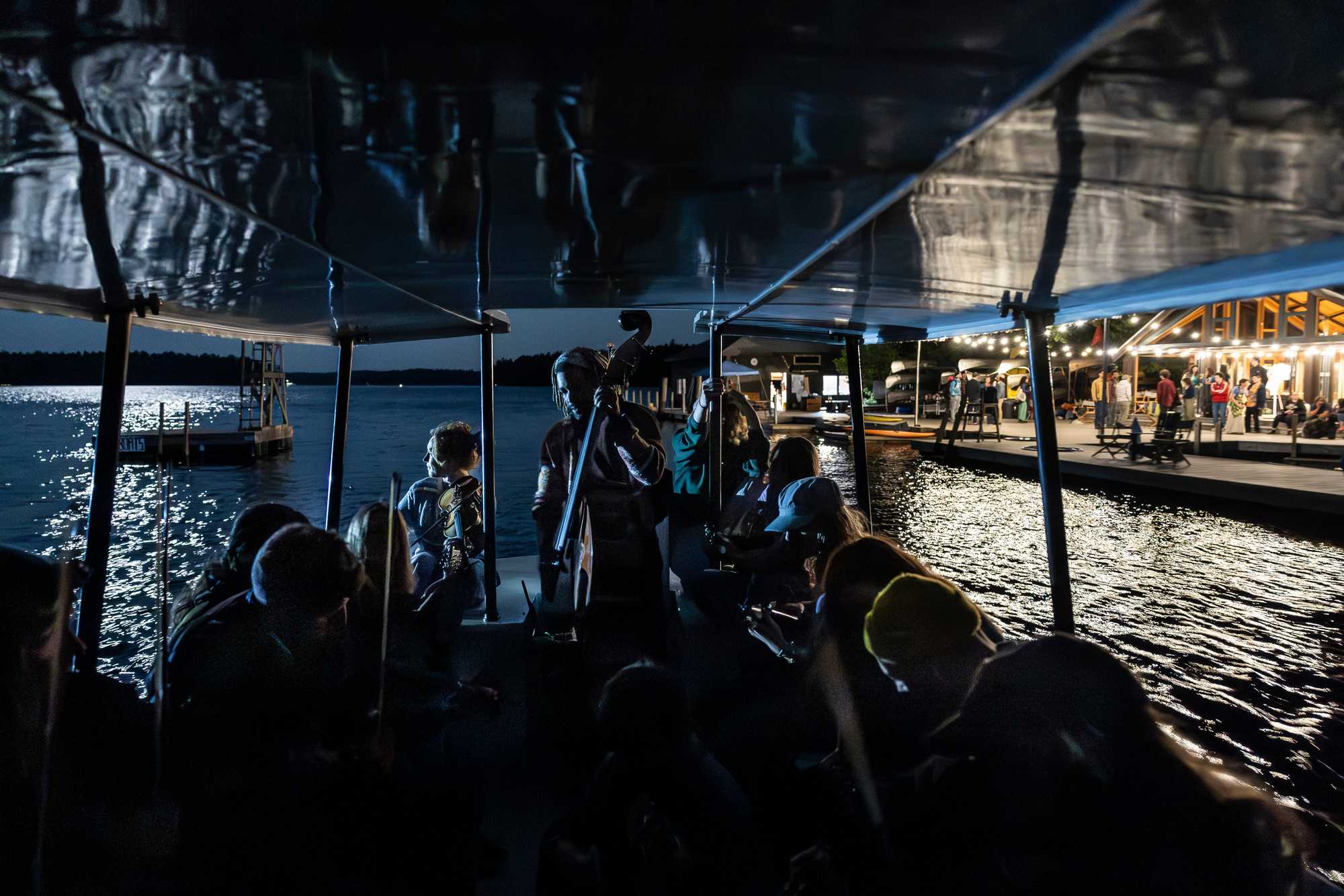 Nelson Williams (back center) plays bass on Dinty Child’s midnight boat ride, passing the camp’s lit-up dock and rec hall on the right. Every year, island manager Child takes campers on late-night excursions under the moon.