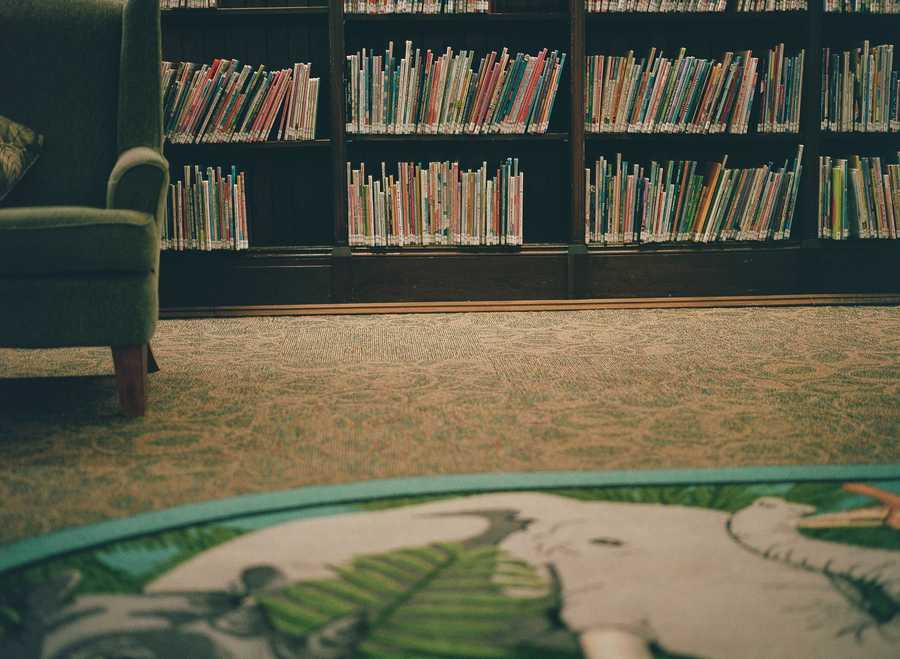 This is the bottom right portion of a composite of four images that make up one image of a green chair in a library. This section contains the bottom right portion of the chair. The chair rests on a textured, patterned carpet. In the foreground is a portion of a rug portraying the head of an elephant. Behind the chair is a wall of bookshelves filled with colorful children’s books.