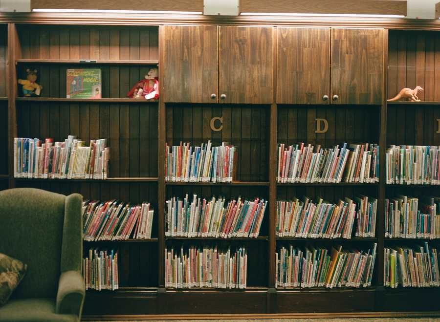 This is the top right portion of a composite of four images that make up one image of a green chair in a library. This section contains the upper right portion of the chair. Behind the chair is a wall of bookshelves filled with colorful children’s books, labeled C and D in this portion of the image. Some of the shelves contain children’s toys, such as dolls and a dinosaur. The book “Goodnight Moon” is given prominence on the uppermost shelf.