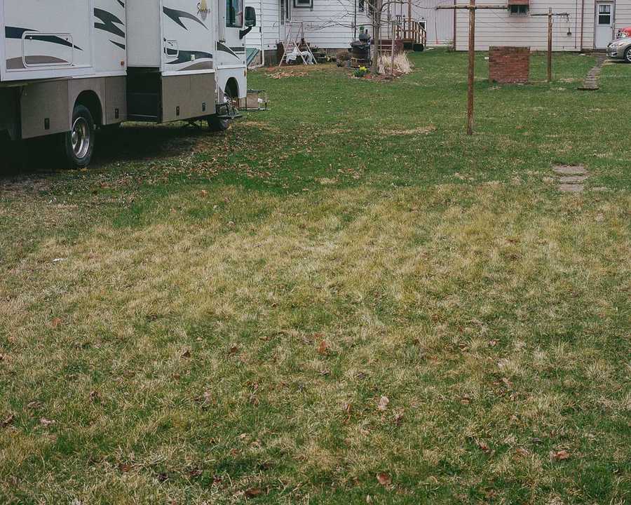 This is the bottom left portion of a composite of four images that make up one image of Kate's childhood home. This portion of the composite shows the lawn splotched with green and yellow grass. A small, concrete path, partially grown over by grass, is barely visible, leading to an empty spot in the lawn where Kate's garage used to be. Two metal posts, capped by a cross-beam, form a clothesline next to the path. A small brick barbecue pit, approximately 3-feet tall, sits beyond the clothesline. The bottom portion of the RV extends out of the top left of the image.