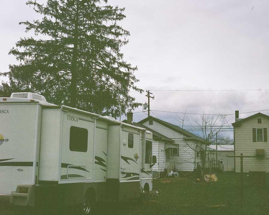 This is the top left portion of a composite of four images that make up one image of Kate's childhood home. This portion of the composite shows a second-story window of the home, framed by dark shutters. To the left is another home, and in the foreground is an RV. Behind the RV towers a solitary tree. Power lines cross above the homes in front of a cloudy, gray sky.