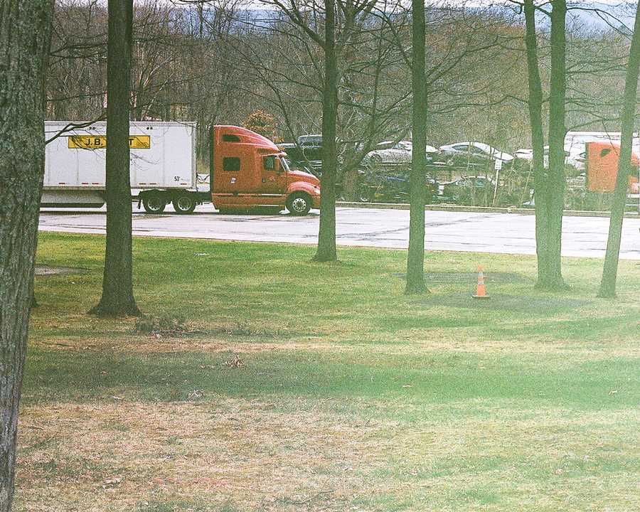 This is the bottom right portion of a composite of four images that make up one image of the rest stop that Kate remembers being taken to as a child. This portion of the composite shows a grass lawn and the base of the trees interrupting a gray sky above. A solitary orange cone rests on the lawn between two trees. Beyond, in the parking lot of the rest stop, is a red semi truck hauling a white trailer. Farther back, another semi hauling a trailer full of cars and trucks.