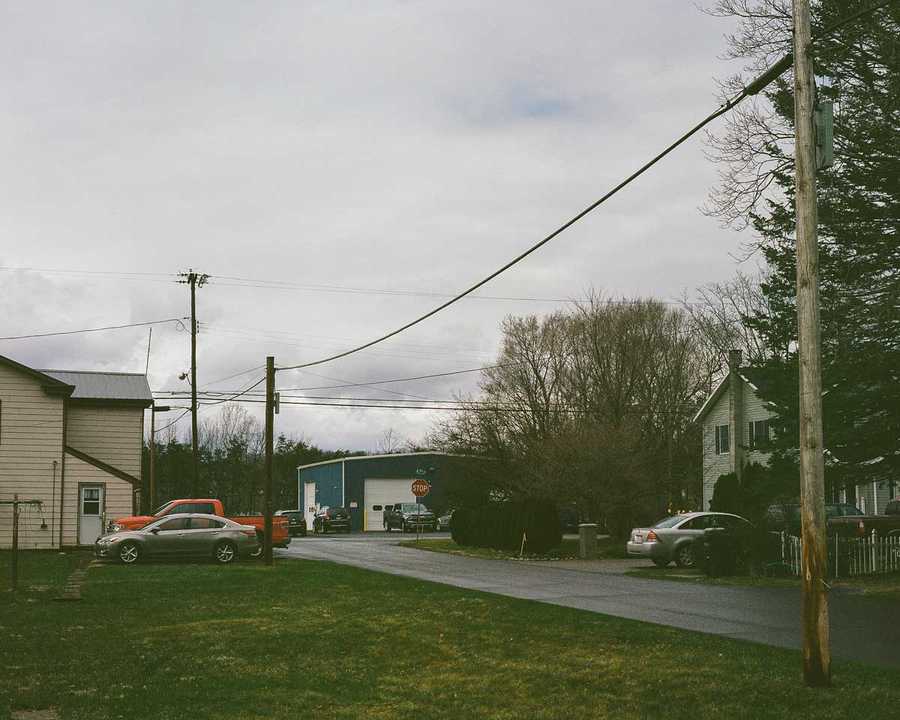 This is the top right portion of a composite of four images that make up one image of Kate's childhood home. This portion of the composite shows the remainder of the upper part of Kate's home. A gray car and red truck are parked in a driveway just off a small paved road that ends in a T-intersection. Across the street is another home, and beyond the intersection is a blue warehouse with large white garage doors. Power lines cross above the homes in front of a cloudy, gray sky.