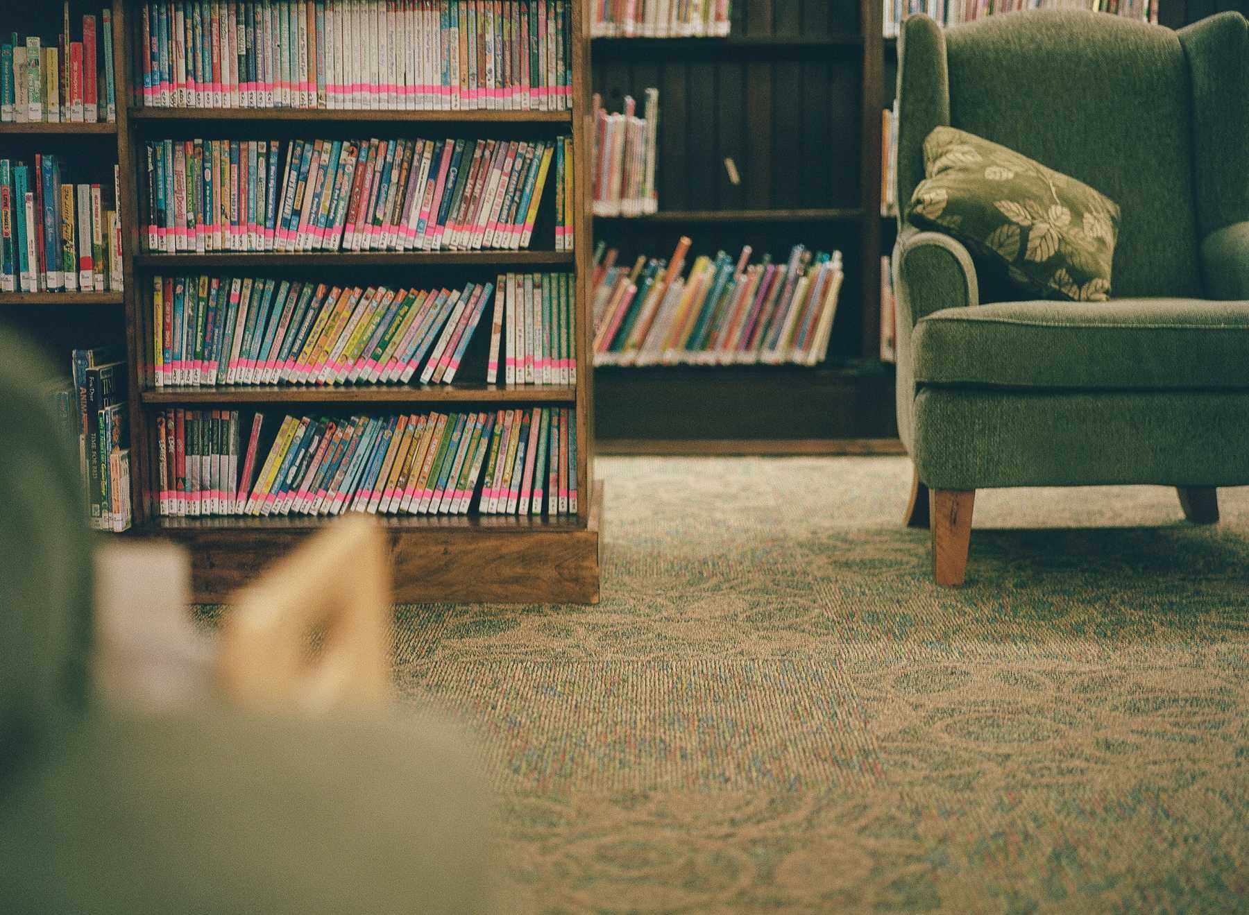 This is the bottom left portion of a composite of four images that make up one image of a green chair in a library. This section contains the bottom left portion of the chair. The chair rests on a textured, patterned carpet. To the left of the chair is a bookshelf packed with books. Each book has a pink sticker on the spine above the white library sticker. In the foreground is a blurred portion of another chair.