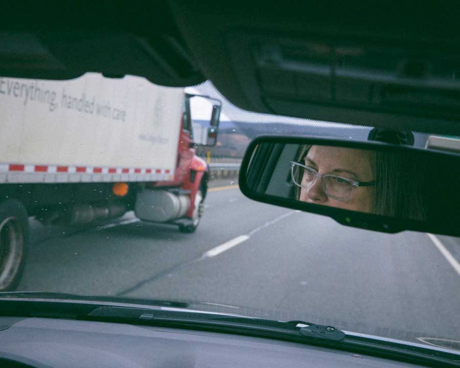 Kate Price’s face is reflected in a rear-view mirror as she drives down the highway. A large truck that reads "Everything handled with care" on the side passes her.