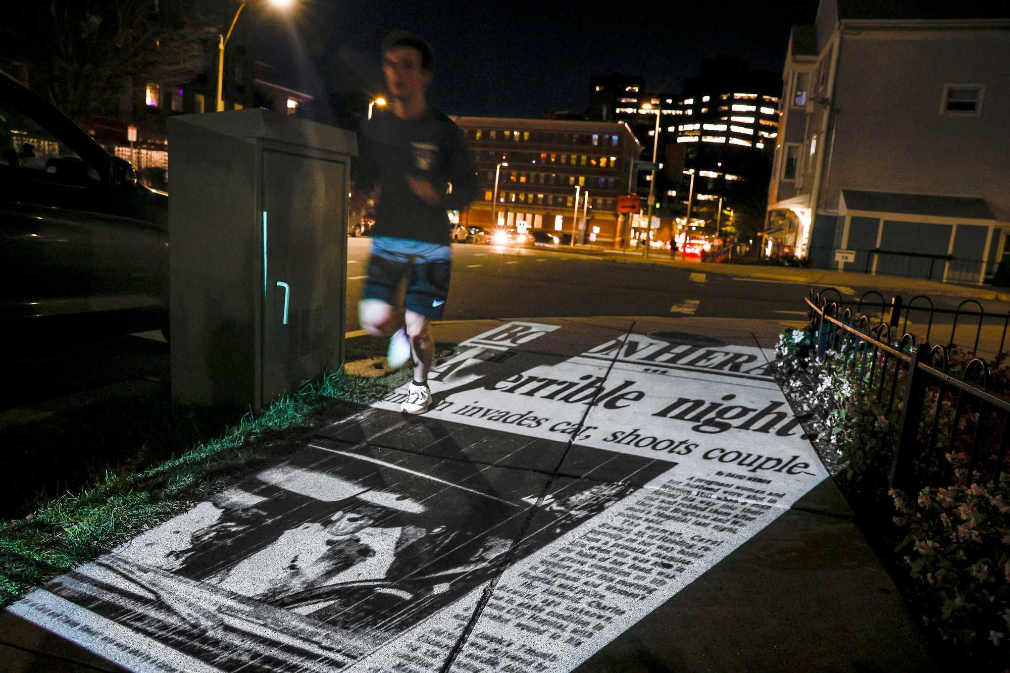 A projection of the Boston Herald’s front page from Oct. 24, 1989, the morning after the Stuart shooting, is cast on the ground as a jogger passes through the intersection of St. Alphonsus Street and Horadan Way. Police found Carol and Charles Stuart shot in their car nearby.