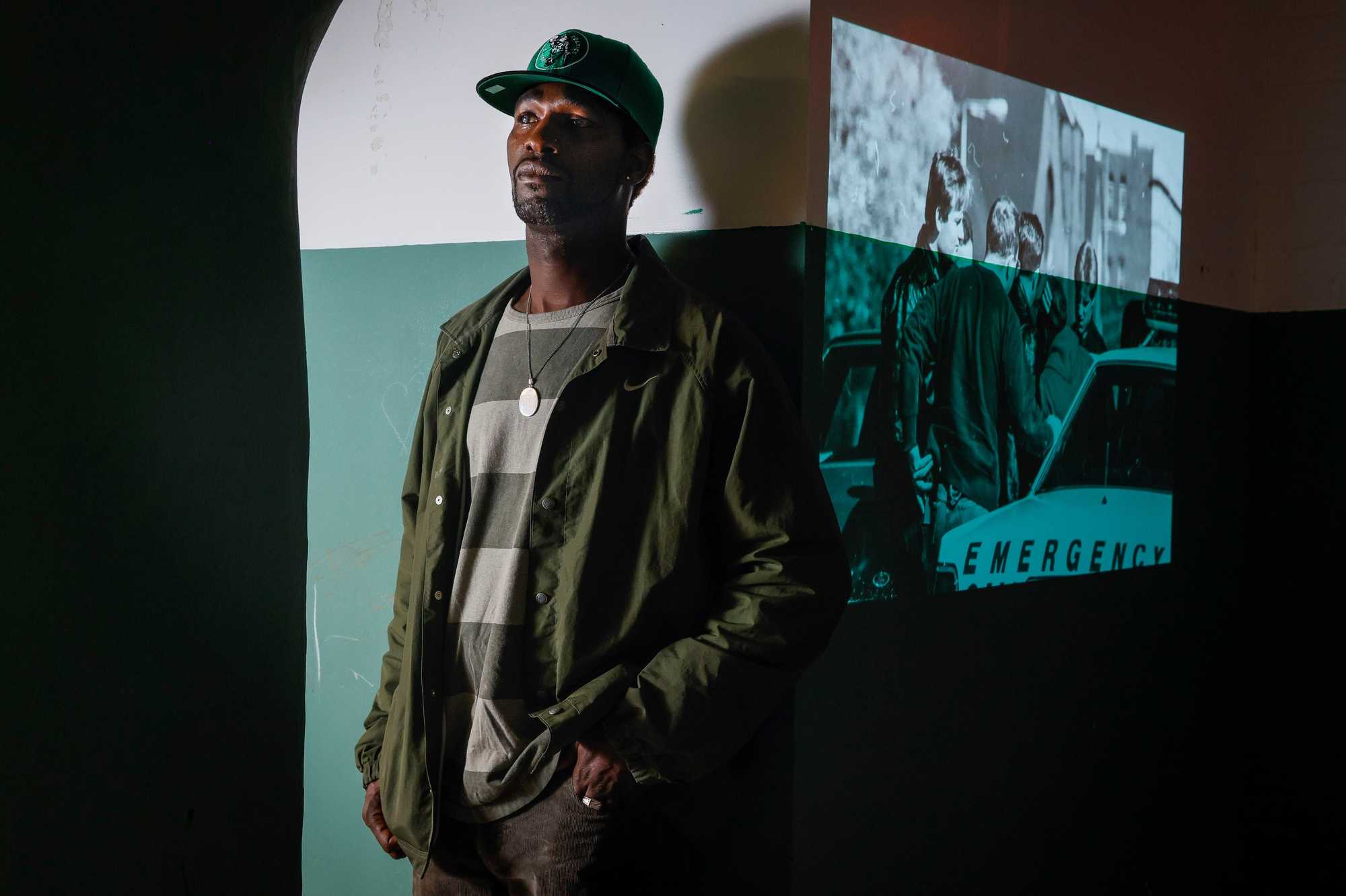 DonJuan Moses, next to a projection of a 1989 image of police officers detaining a man in Mission Hill. Moses was 11 years old when police burst into his family's home and arrested his cousin. 