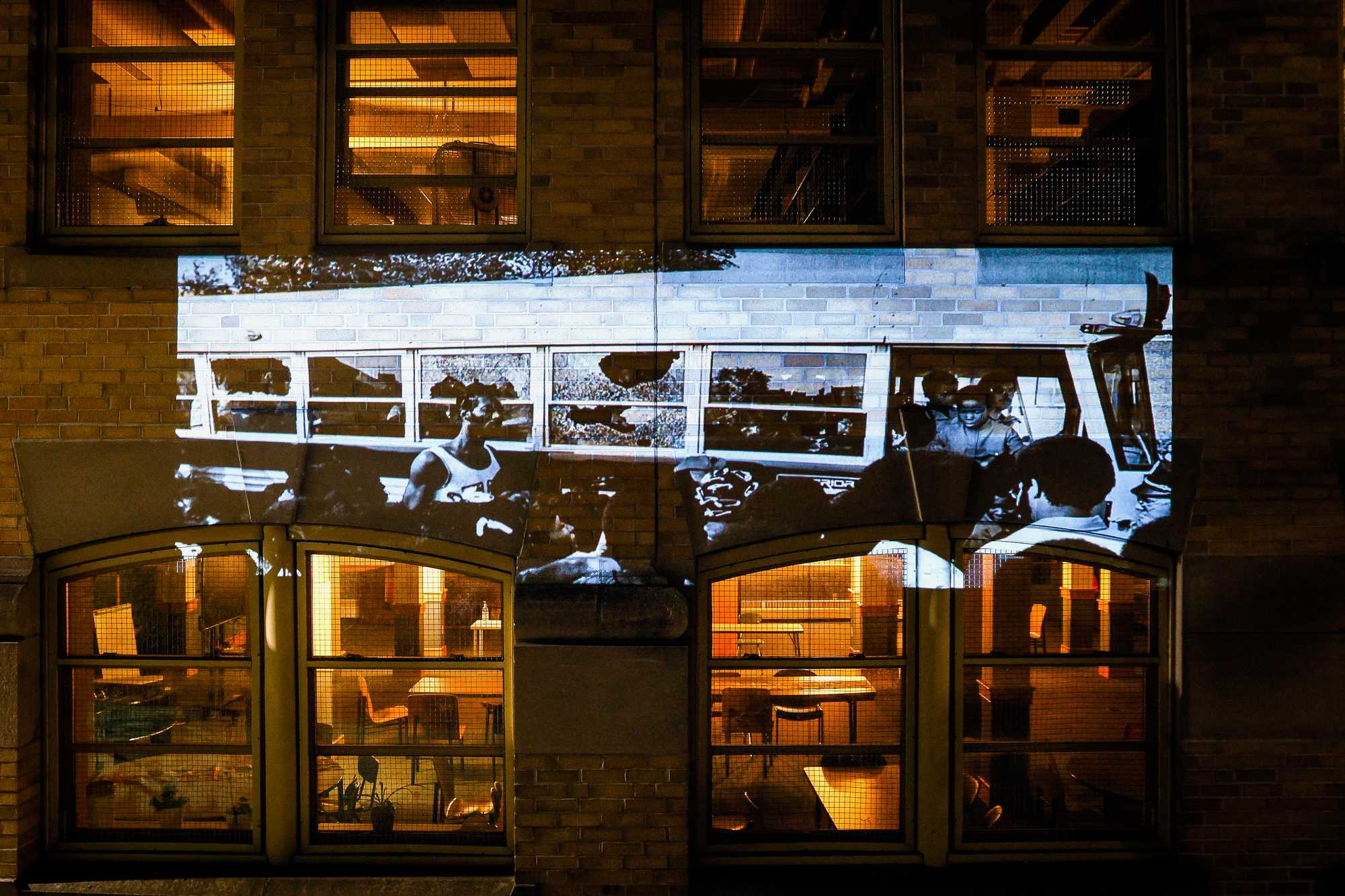 A photograph of a bus with broken windows, carrying students as it returned to Columbia Point on Sept. 12, 1974, is projected on the former South Boston High School.