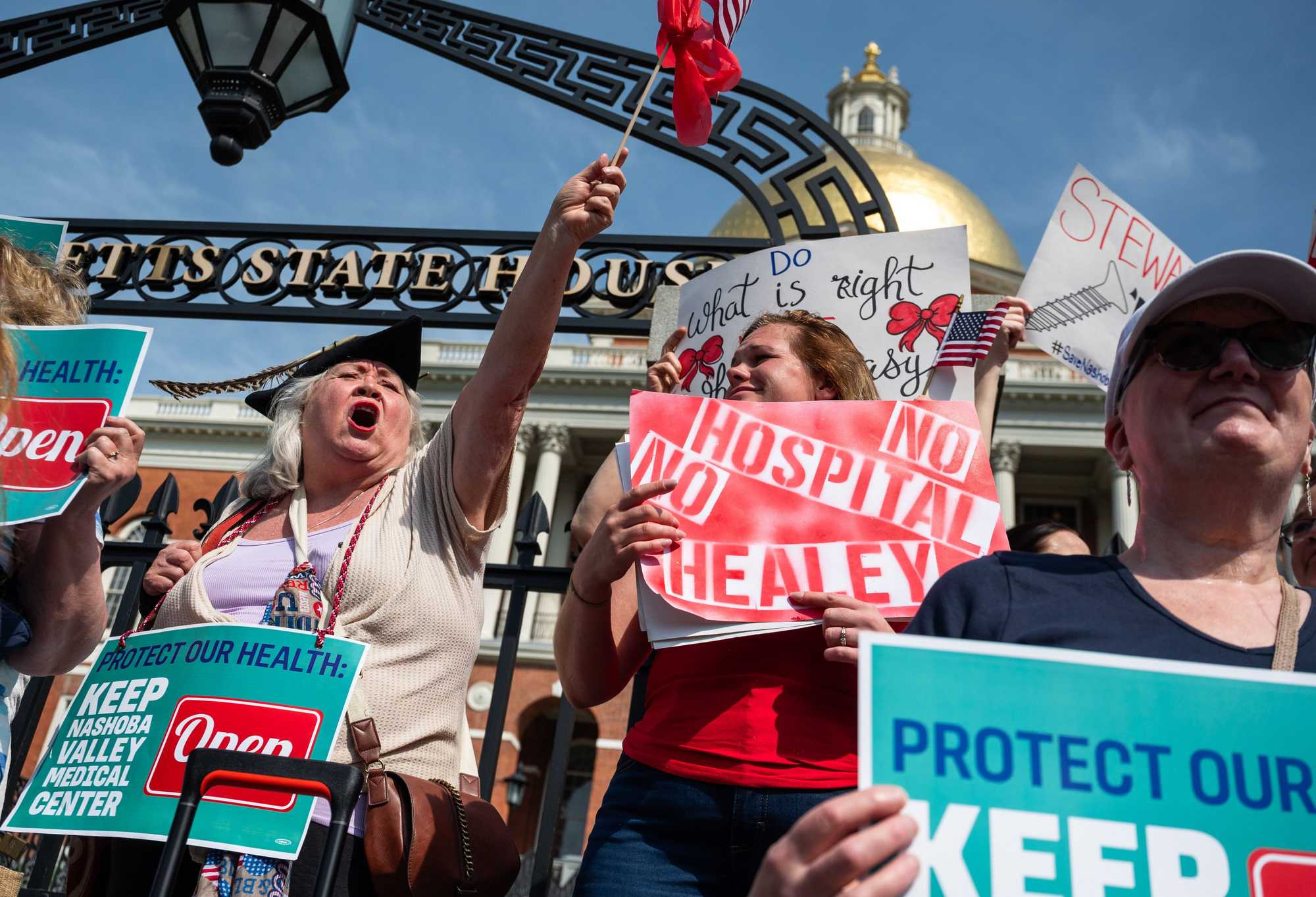 Eleanor Gavazzi (left, waving flag) shouted at a rally advocating to keep Steward hospitals Carney Hospital and Nashoba Valley Medical Center open on the front steps of the State House in Boston on Aug. 28. Gavazzi said that Nashoba Valley Medical Center saved her son's and mother’s lives and that it would take her 40 minutes to get to an ER if it  closed.