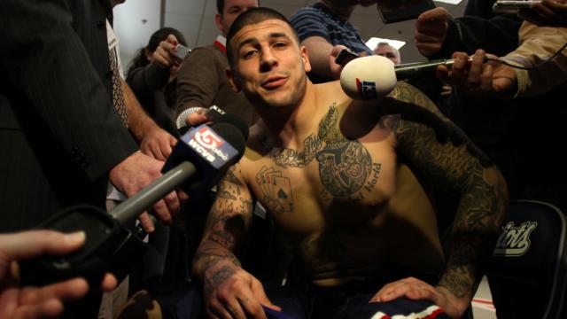A shirtless Aaron Hernandez sits on a bench with several television camera microphones pointed at his face